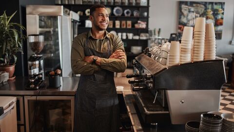 Ein lachender Mann mit verschränkten Armen steht hinter dem Thresen seines Cafes