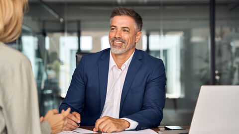 Mann im Anzug sitzt am Schreibtisch gegenüber einer Frau und lächelt